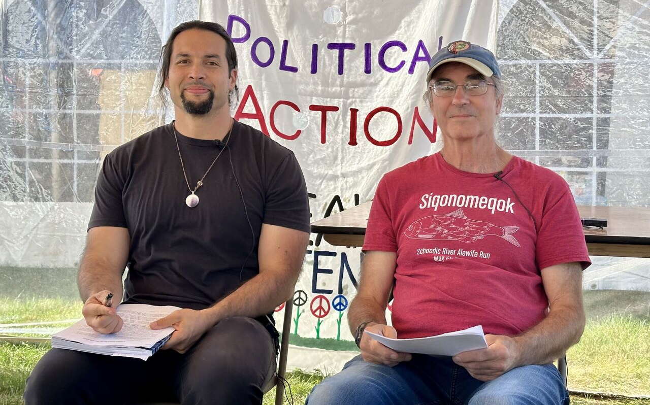 Two people sit with documents under a tent.