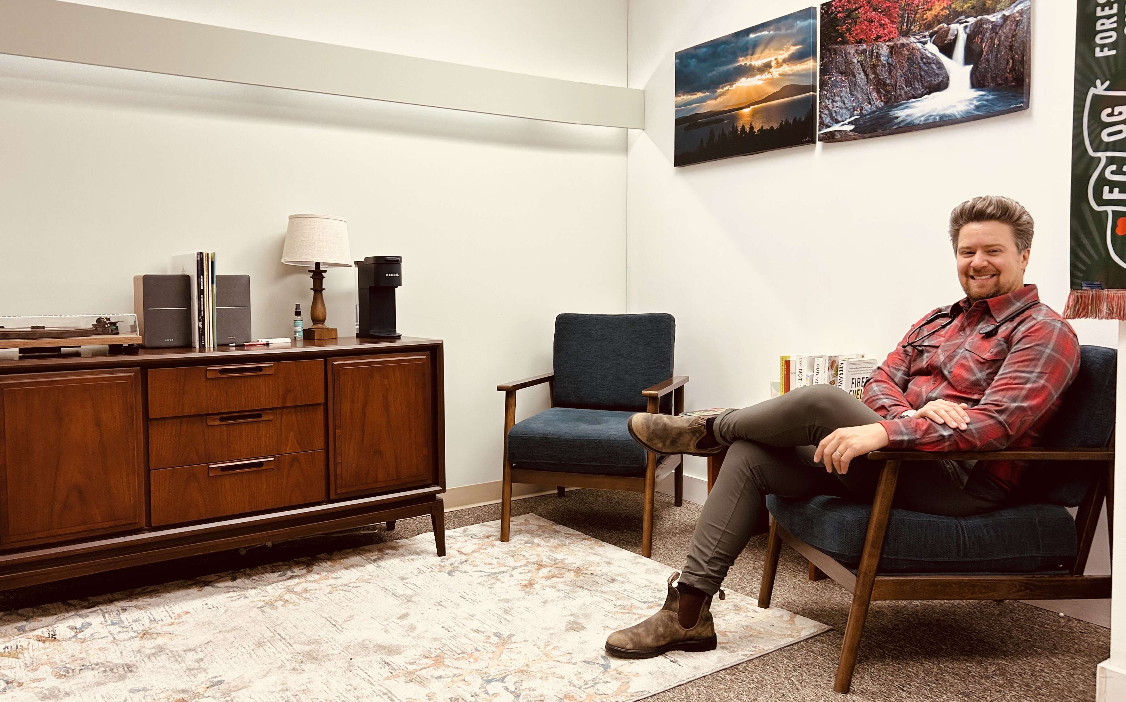 A person sits in a room that has chairs and a sideboard.