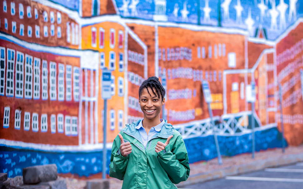 black woman with her thumbs up, in front of a mural
