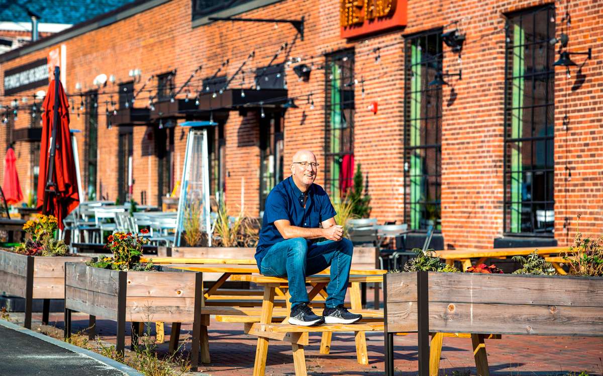 A smiling white man sits on a picnic table on a brick patio in front of a long brick building with neon signs