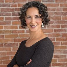 A woman with a dark complexion and curly black hair stands in front of a brick wall with arms crossed smiling at the camera