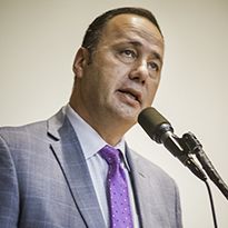 A man wearing a gray suit and purple tie speaks into a microphone