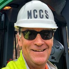 A smiling white man in sunglasses with a hardhat that says MCCS and yellow raincoat in front of heavy equipment