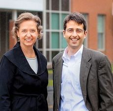 A smiling woman and man in business attire stand in front of a brick building