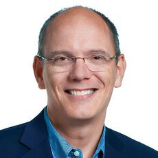 A head and shoulders photo of a smiling white man with glasses in a boue suit jacket and blue shirt