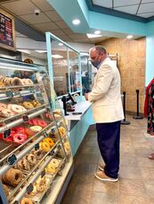 A man, white, wearing a face covering and dressed in a suit, in front of a glass case showing a variety of doughnuts he looks like he's trying to decide which to buy