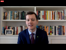 screenshot of man in suit and tie sitting in front of bookcase