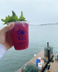 Hand holding a cocktail in a plastic cup over water and a pier with lobster traps