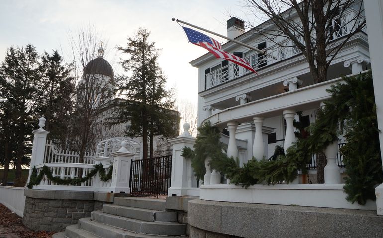A closeup view of the front of a Georgian-style white house with a curved white fence in front, an American flag flying from the porch roof, granite steps leading up from a brick sidewalk and Christmas punting on the fence, with the Maine State House, a large granite building with a copper dome, in the background.
