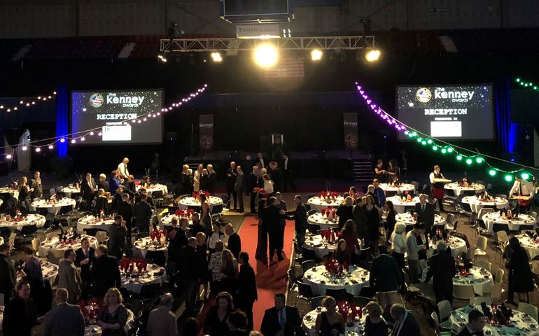 A banquet room with many people sitting at round tables and a big screen that says Kenney Awards on it.