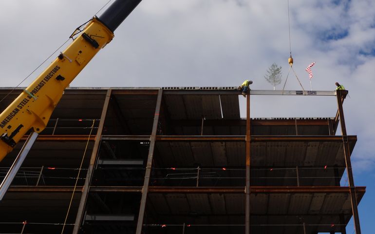 A crane lifts an evergreen and American flag to the top of a steel frame for a strucutre as two workers on a girder steady it.