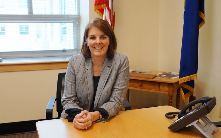 A woman sits at a desk, smiling at the camera, with an American and Maine flag behind her