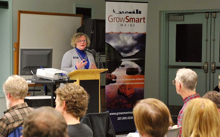 A woman stands at a podium, speaking to a seated audience with a banner next to her that says GrowSmart Maine