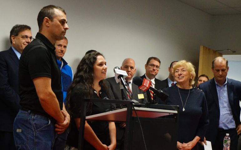 A couple speaks at a lecturn with microphones on it as Maine Gov. Janet Mills smiles in the background