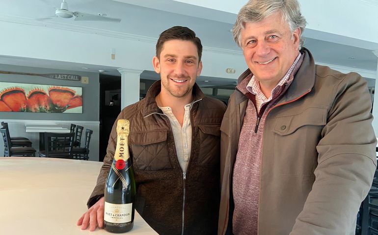 A young man and older man at a counter with a bottle of champagin smiling for the camera