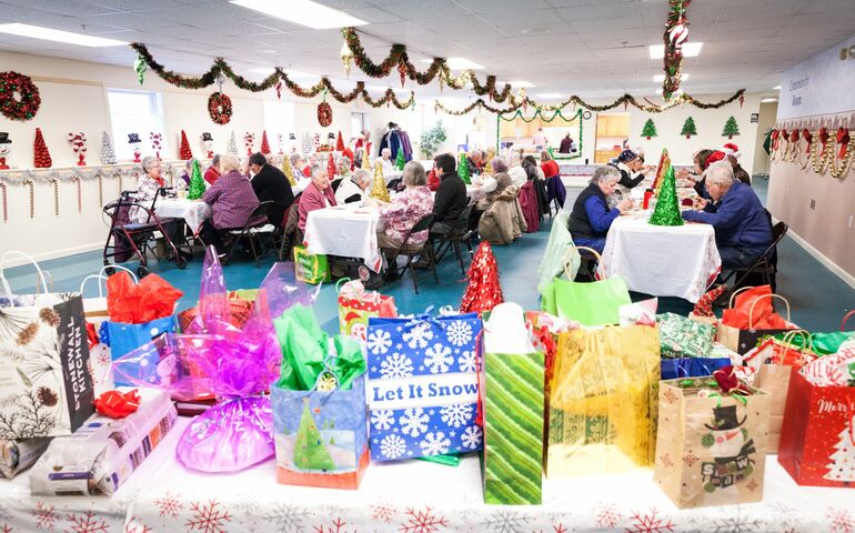 Seniors at a holiday gathering -- gift table in foreground, people at tables eating in the background.