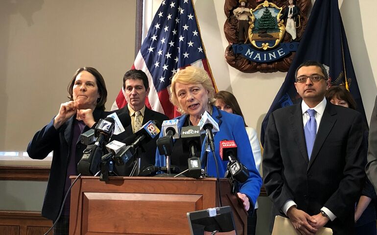 Gov. Janet Mills stands behind a lecturn with a state of Maine seal and several people standing behind her