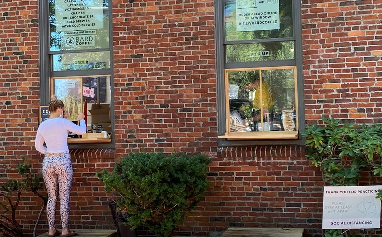 A woman wearing a protective face covering buys a coffee at a walk-up window in a brick building while a sign nearby thanks customers for practicing social distancing
