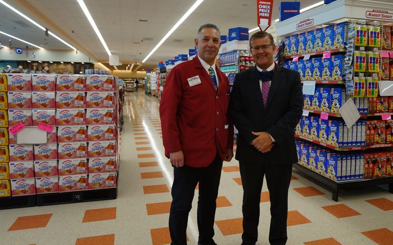 Two men standing in the aisle of a large supermarket, one with a red blazer on it that says Market Basket
