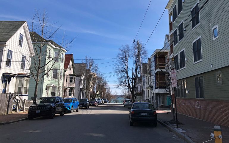 A narrow city street with three-story wooden apartment buildings on the left and a large modern condominium development on the right, at the end of the street the ocean can be seen