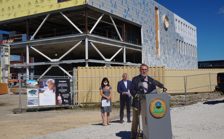 A man stands outside at a lectern on a sunny day with an underconstruction building behind him