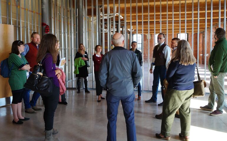 A group stands in a partially finished industrial looking building, as a tall man in a vest talks to them