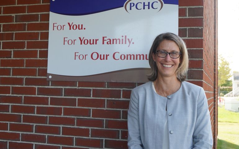 a smiling white woman with short blond hair and classes stands in front of a brick wall with a sign on it that says PCHC for you for your family for our community
