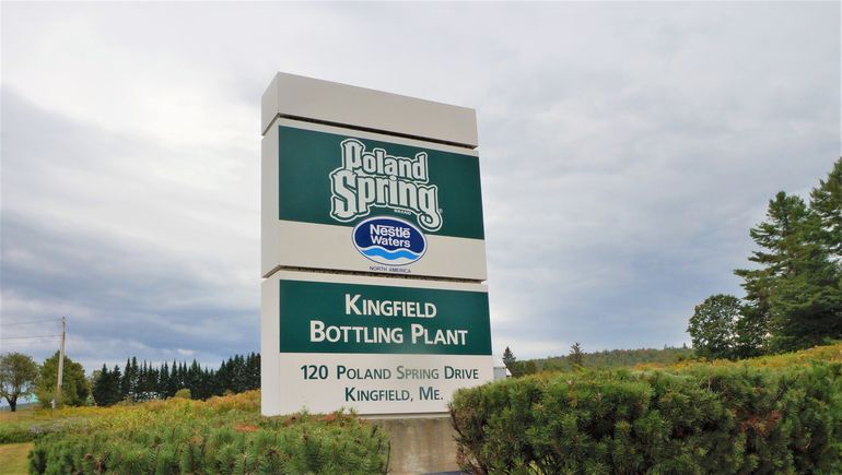 A sign that says Poland Spring Nestle Waters Kingfield Bottling Plant with rural fields surrounding it, and pine trees in the background