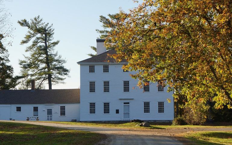 A colonial three-story white clapboard building in autumn, with a tree showing bright foliage next to it