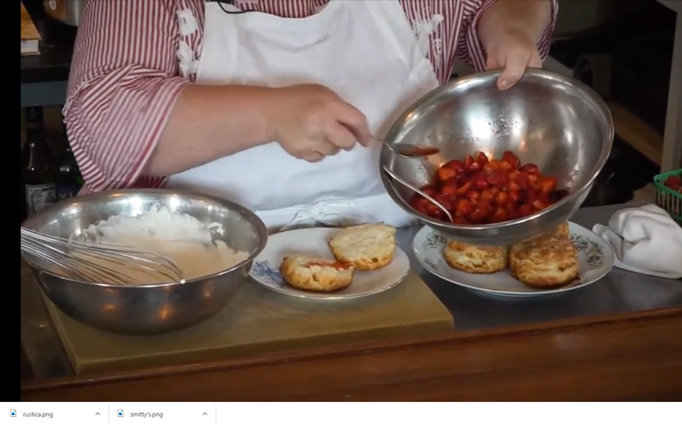 a woman with a large bowl of strawberries is tipping it to ladel them onto freshly made biscuits as a large bowl of whipped cream stands by