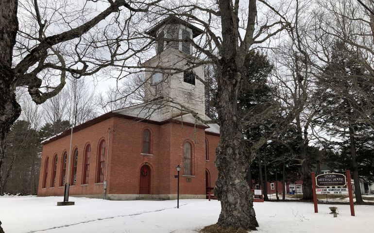 An old brick building with a steeple with white peeling paint and a snow-covered yard