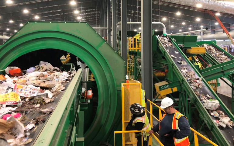 the interior of a large building with a lot of conveyor belts, including a closeup of one with garbage and a man with a hardhat on a ladder 