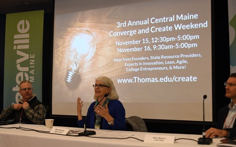 A woman sitting at a table with a screen behind her projecting an image that says thomas college converge and create conference speaks into a microphone