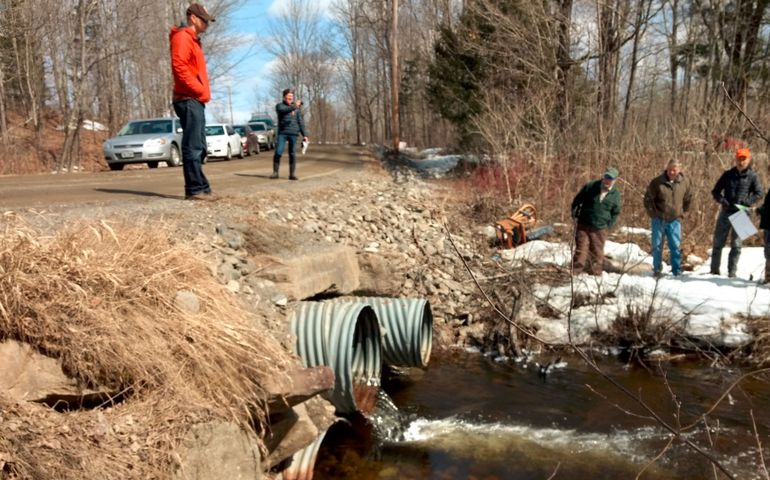 Several men stand on a road overlooking some old culverts discharging water into a brook, it's winter and there's snow on the ground.