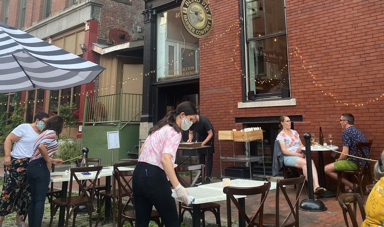 Servers wearing masks clean tables as customes eat at a sidewalk eating area in portland next to a brick building