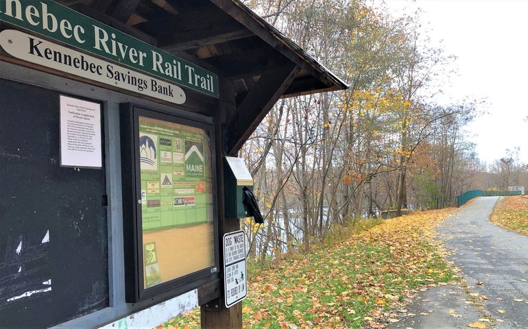 A kiosk that says Kennebec River rail trail, with a paved trail winding through leaf-covered grass and a river off to the left and train tracks to the right