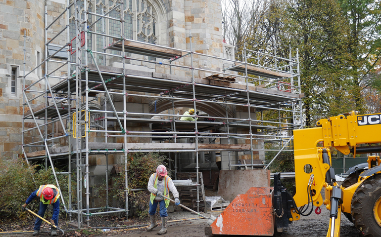 two men wrok with congrete next to a stone shapel with scaffolding on it as a heavy equipment frontloader stands by