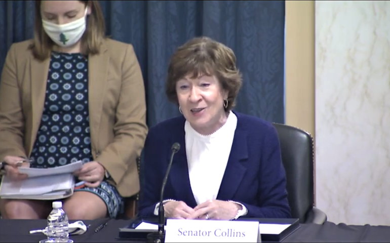 A woman in a purple suit and white blouse at a table with a sign that says senator collins in front of her talks