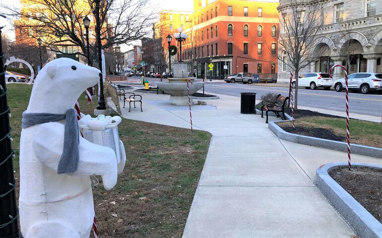 A polar bear statue holding a present stands next to a sidwalk lined with candy canes in a small park as brick buildings of a downtown are in the background