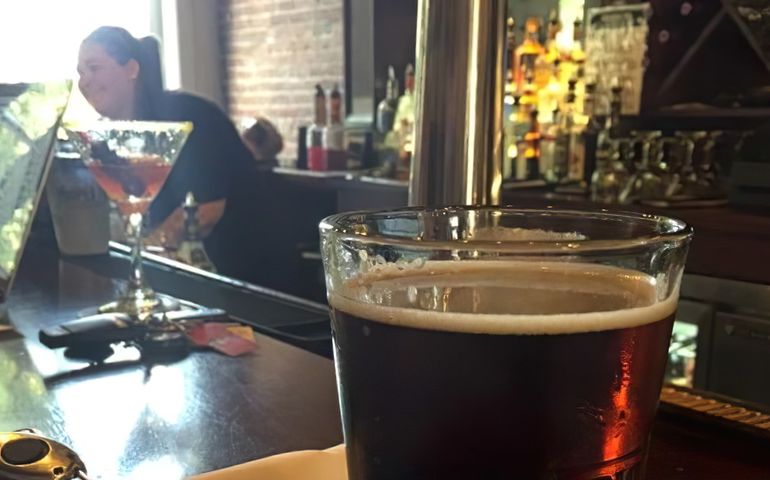 a bar with a closeup of a dark beer and an out of focus image in the background of a bartender and a martini like drink