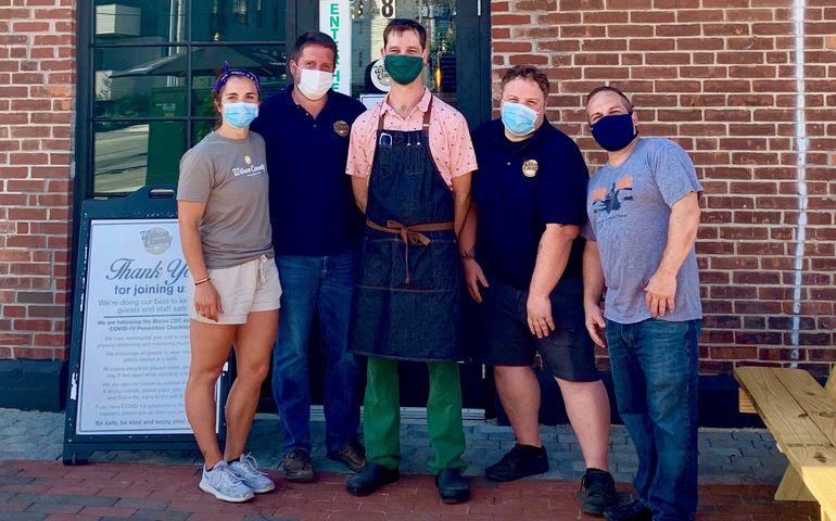 A group of restuarant workers, wearing face coverings, stands on a brick sidewalk in front of their restuarant