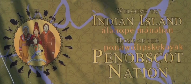 A green sign says welcome to indian island home of the penobscot nation with a logo showing a native family and a backtround of a river running across a map it also has the words in native writing