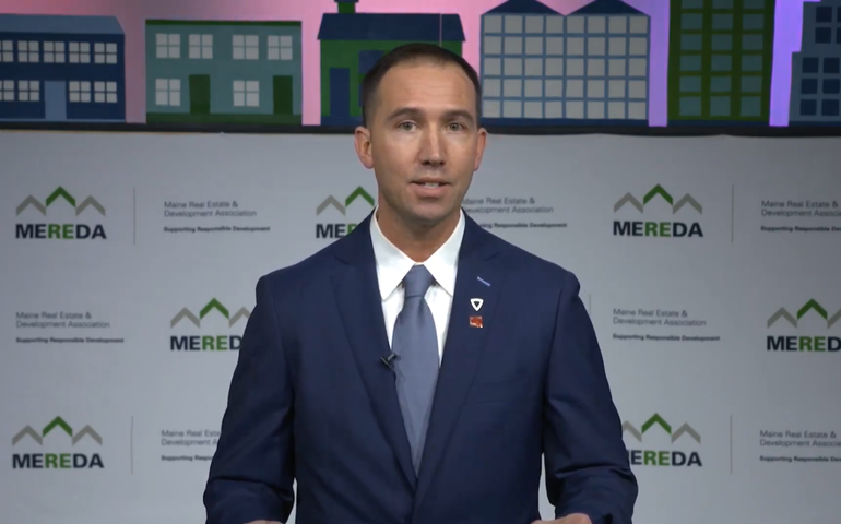 A man in a suit talks in front of a background that says MEREDA