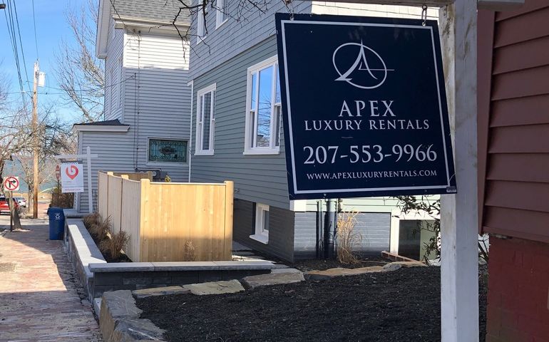 a closeup of a sign that says apex luxury rentals next to a clapboard building and a view down the street to portland's eastern promenade