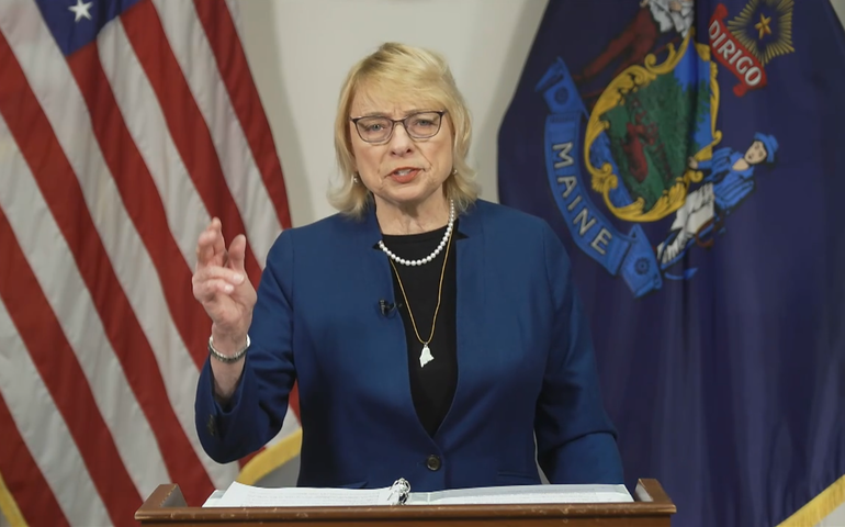 A woman in a navy blue suit and short blond hair speaks at a lecturn in front of an american flag and a maine flag she looks serious and is gesturing with her right hand