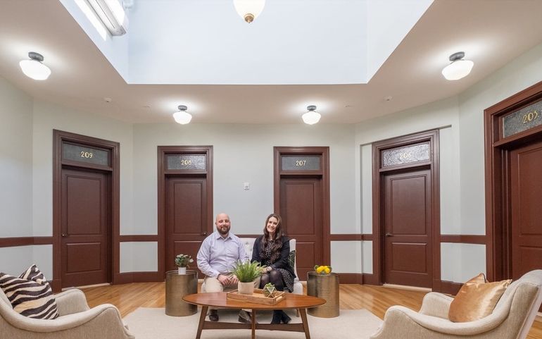 A manand woman sit on a small couch behind a coffee table with old wood doors on the walls behind them