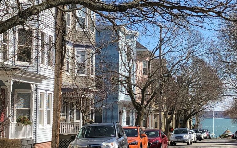 A row of pastel-colored triple deckers close to the street with the blue of the ocean at the end of the street and the trees are bare