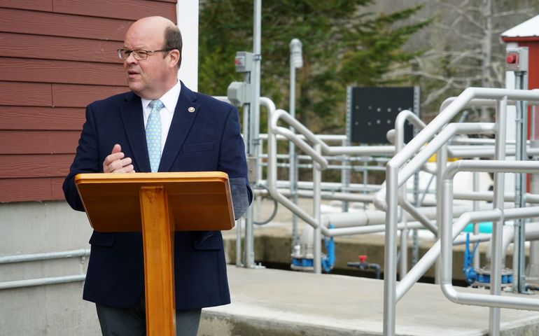 A man stands at a lecturn with a maze of piping behind him