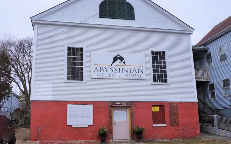 A three story clapboard building with the first story bright red break and a peaked roof with a large sign across the front that says abyssinian meeting house