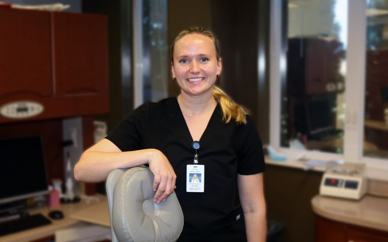 A smiling white woman in dark hospital srubs leans on a dental chair